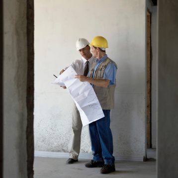 Two construction workers in hard hats review blueprints inside an unfinished building. The setting is calm, with a focus on planning and collaboration.