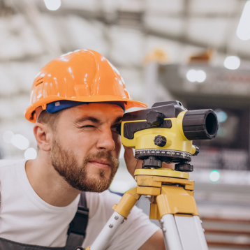 A surveyor in an orange hard hat looks through a theodolite on a tripod in an industrial setting, focusing intensely. Background is blurred.