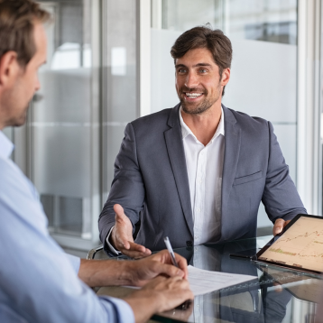 An office meeting with two men. One, in a suit, smiles while holding a tablet, gesturing. The other writes notes, suggesting a collaborative atmosphere.