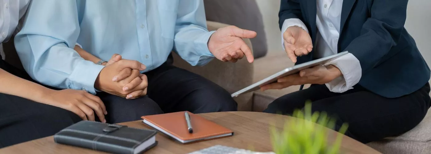 Three people sit at a table, discussing documents with notebooks and a tablet. They appear engaged and attentive, suggesting a professional meeting.