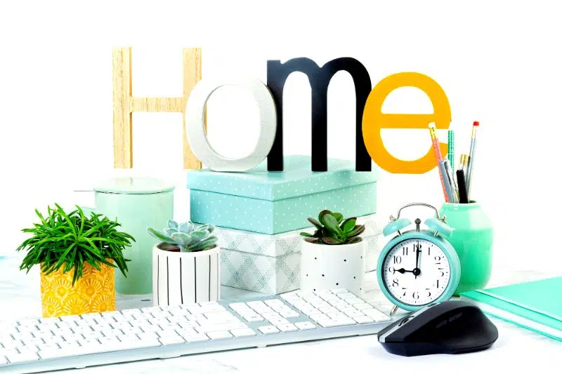 A bright, organized desk setup with "Home" in colorful letters. Features a keyboard, clock, mouse, plants, boxes, and pencil holder, conveying a cheerful and tidy workspace.
