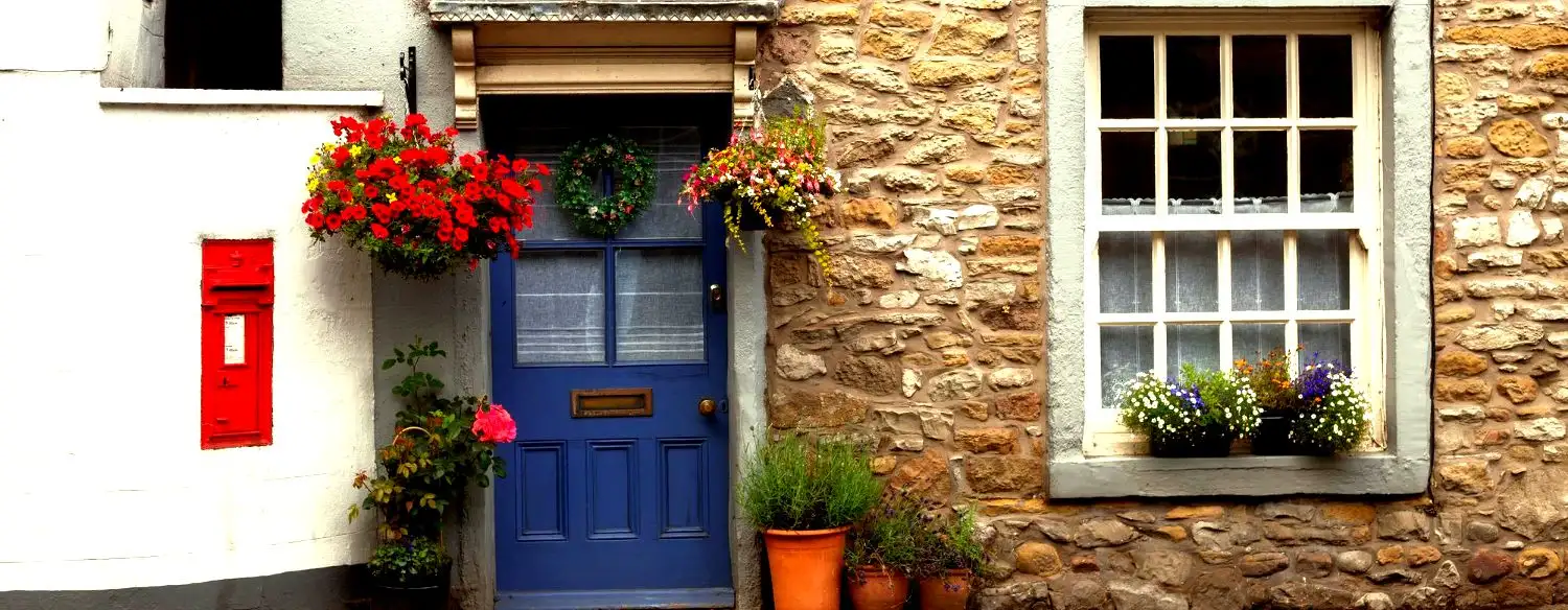 Charming cottage with a blue door, vibrant red flowers above, and potted plants. A red mailbox on the white wall adds a pop of color. Cozy and inviting.