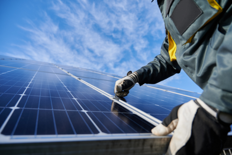 A person wearing gloves and a work jacket adjusts a solar panel under a bright blue sky. The scene conveys a sense of focus and sustainable energy effort.