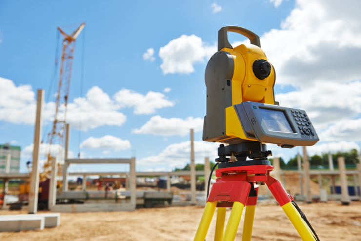 Yellow surveying equipment on a tripod at a construction site. In the background, a crane and building structures under a bright blue sky.
