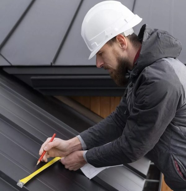 A person in a hard hat and jacket measures a metal roof with a tape measure, focusing intently while writing notes. The scene conveys precision and diligence.